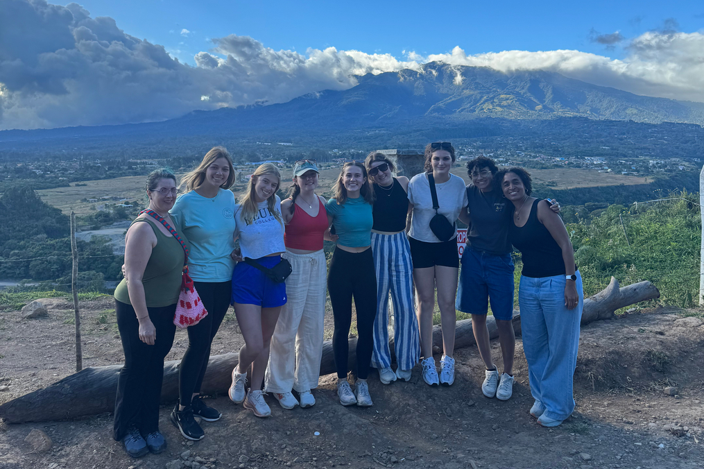 Group photo of eight people standing on a mountain overlook with dramatic clouds and a valley landscape behind them.