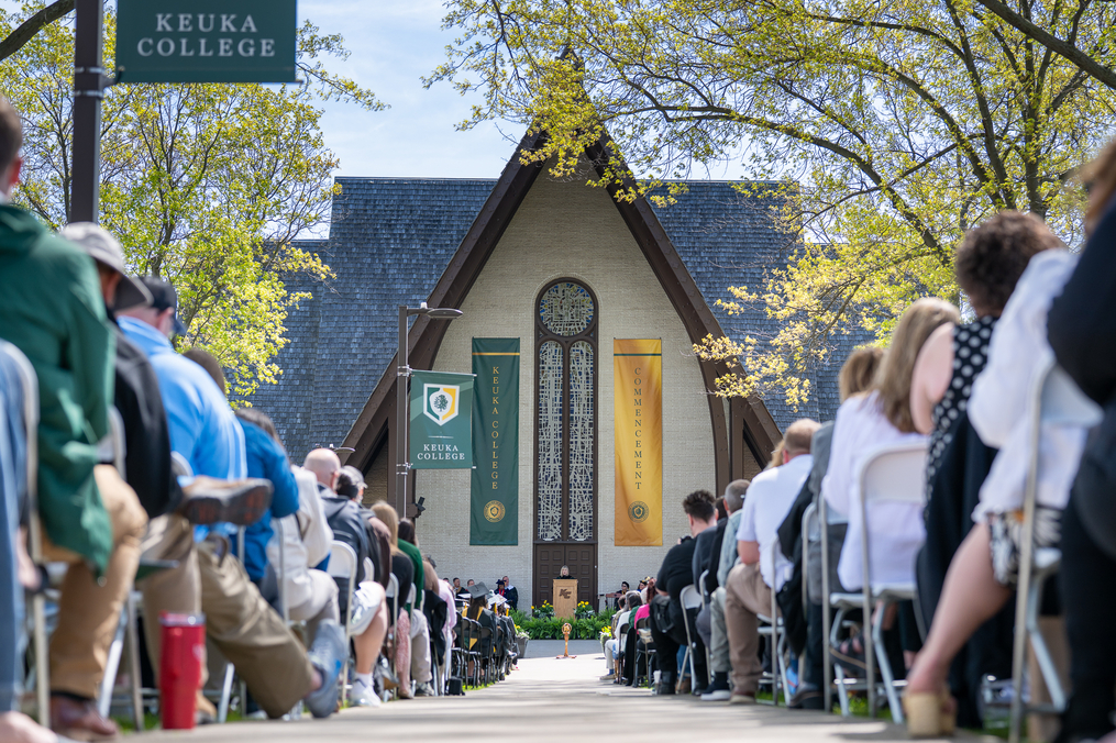 Wide view of Keuka College Commencement ceremony with graduates, families, and faculty seated outdoors facing the campus chapel stage on a sunny spring day.