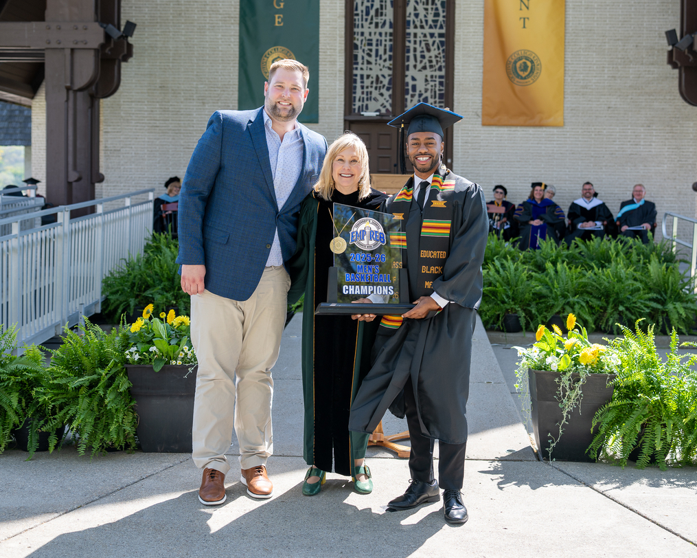 A graduate in cap and gown stands on stage at Keuka College holding a championship trophy beside two faculty and staff members during Commencement ceremonies.